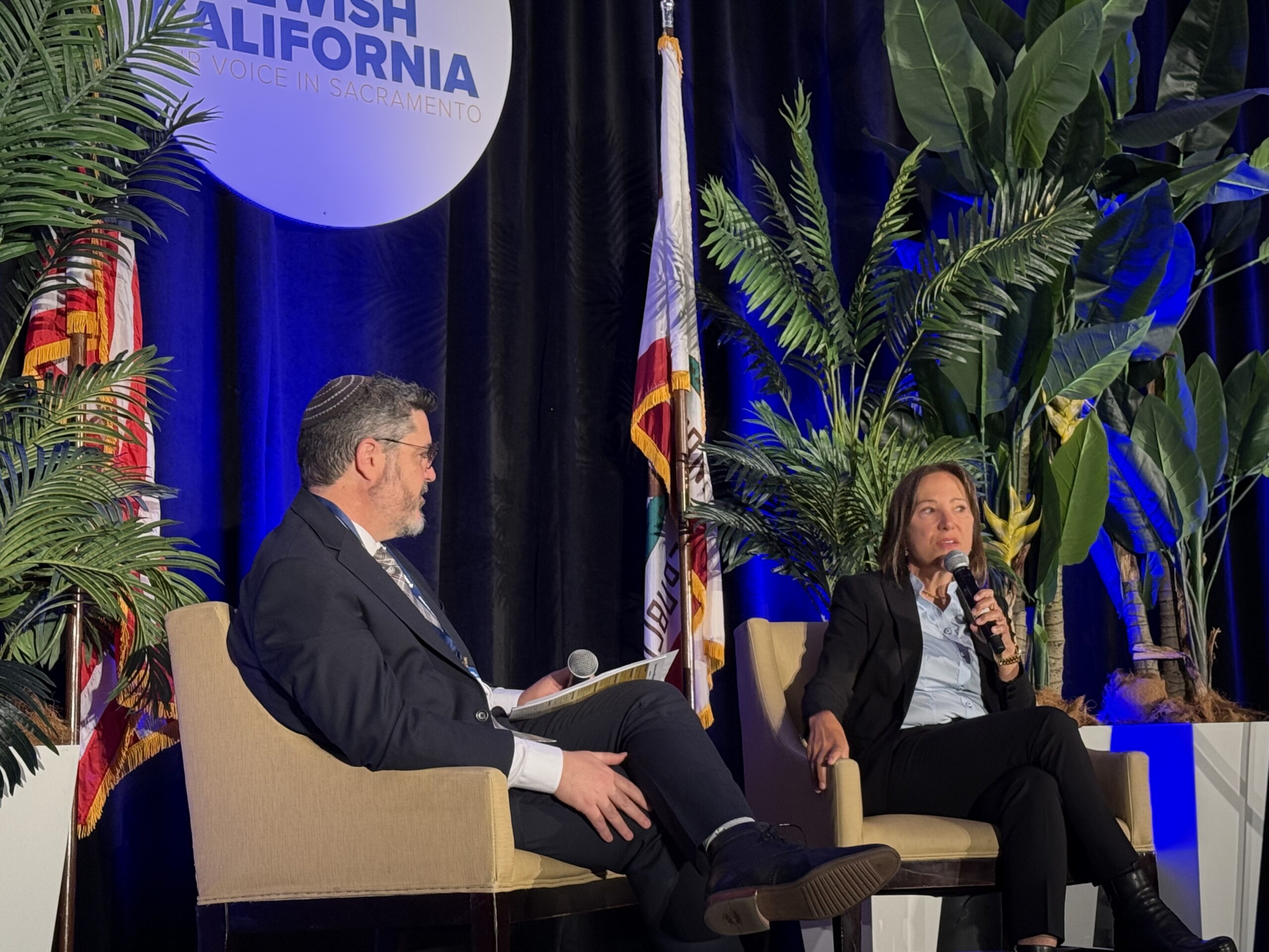Image of Lt. Gov. Kounalakis sitting on the stage with Rabbi Noah Farkas speaking at the Jewish CA event in Sacrament, CA.